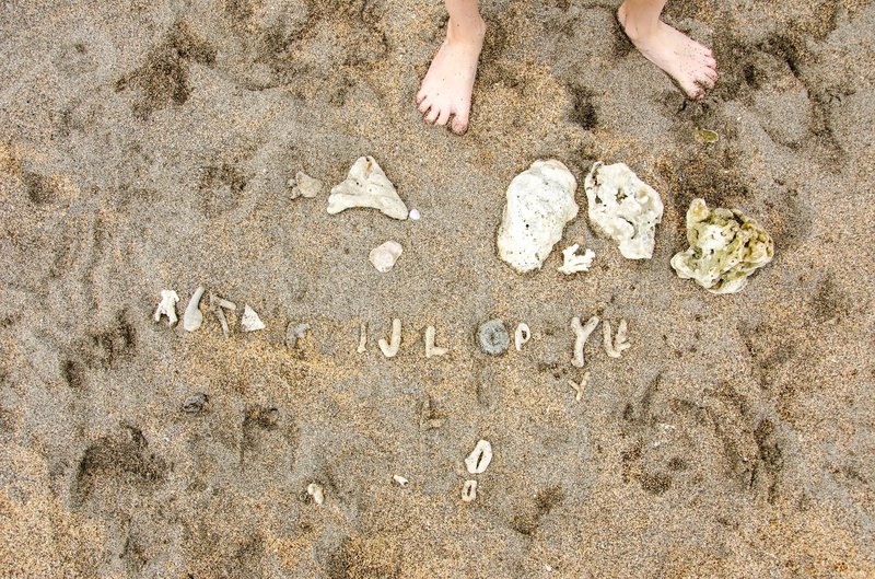 Feet in the sand above scattered pieces of coral arranged like letters of an alphabet.